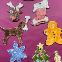 Load image into Gallery viewer, A collection of handcrafted, painted ceramic Christmas ornaments arranged on a pink paper backing over a wooden table. The ornaments include a gingerbread man, a stocking, a Christmas tree, a reindeer, a dog, a snowman, and a snowflake, each with unique hand-painted colors and textures.
