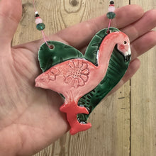 Load image into Gallery viewer, Hand holding a decorative ceramic flamingo on a heart decoration on a wooden surface