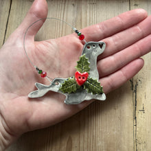 Load image into Gallery viewer, Hand holding a decorative seal-shaped ornament with holly leaves and berries on a wooden surface