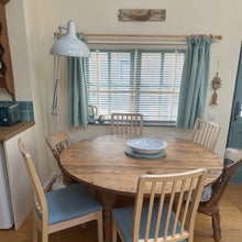 Load image into Gallery viewer, Dining area with wooden table and chairs in a room with large windows and curtains.