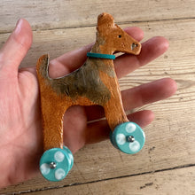 Load image into Gallery viewer, A hand holds a ceramic dog with turquoise and white painted wheels and a turquoise collar, set against a rustic wooden plank background.
