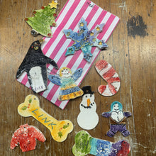 Load image into Gallery viewer, A collection of hand-painted ceramic Christmas ornaments and a pink striped gift box arranged on a worn wooden tabletop. The ornaments include a snowman, a candy cane, a stocking, a penguin, a Christmas tree, and a bone ornament