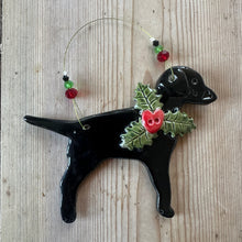 Load image into Gallery viewer, A black ceramic Christmas ornament shaped like a labrador wearing a green holly wreath with red berries, displayed on a light-colorued wooden surface.
