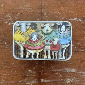 Small rectangular tin with illustrations of sheep wearing colorful sweaters on a wooden surface.
