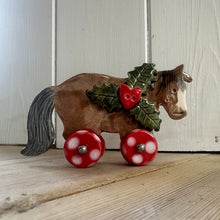 Load image into Gallery viewer, Horse with red wheels and holly decorations on a wooden surface
