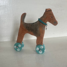 Load image into Gallery viewer, A ceramic  dog figurine with a brown and black body, wearing a teal collar and teal p&amp; white spotty wheels, displayed on a white shelf.
