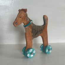 Load image into Gallery viewer, A small ceramic dog with turquoise wheels and a turquoise collar, standing on a white shelf against a white wall.
