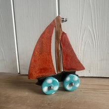 Load image into Gallery viewer, Small sailboat model with a red sail and blue wheels on a wooden surface.
