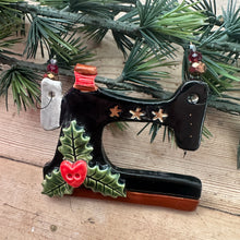 Load image into Gallery viewer, A vintage Christmas ornament shaped like a vintage sewing machine decorated with a glossy black body, gold stars, green holly leaves, and red berries, displayed on a wooden surface next to a green pine branch.
