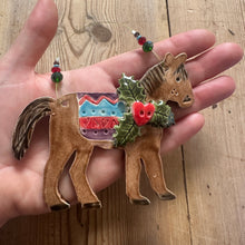 Load image into Gallery viewer, Decorative horse ornament with holly leaves and berries held in a hand on a wooden surface.
