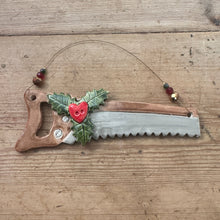 Load image into Gallery viewer, A Christmas-themed saw ornament with a brown handle, silver blade, green holly leaves, and red berries, hanging on a rustic wooden plank background.
