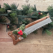 Load image into Gallery viewer, A Christmas-themed saw with a brown wooden handle decorated with green holly leaves and red berries, shown on a light wood plank background next to a pine garland.
