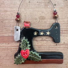 Load image into Gallery viewer, A ceramic Christmas ornament shaped like a  vintage sewing machine featuring a glossy black body with gold stars, green holly leaves, and red berries, hanging from a wire with beads on a wooden surface.
