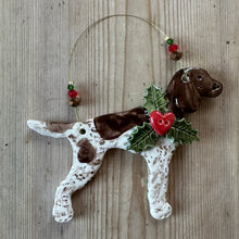 Load image into Gallery viewer, A ceramic Christmas ornament shaped like a white and brown spotted dog  and a green holly collar with a red berry accent, hanging from a thin wire loop on a rustic wooden surface.
