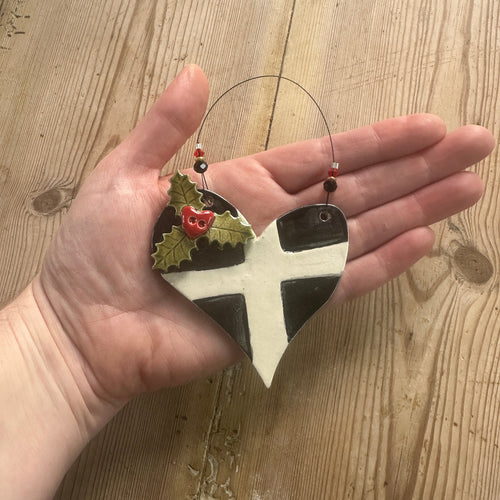 Hand holding a decorative Christmas ornament with a St Piran's flag design on a wooden surface